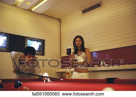 Stock Image - Young man playing pool, woman holding glass of beer and watching. Fotosearch