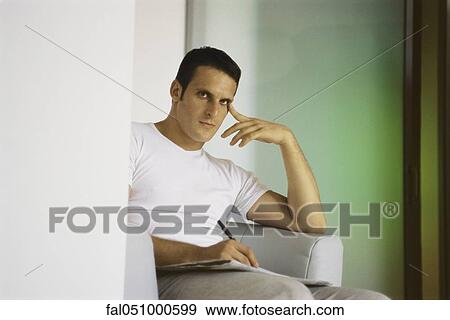 Young man sitting in armchair, holding pen and newspaper, looking at camera View Large Photo Image Stock Photo - Young man sitting in armchair, holding pen and newspaper, looking at camera. Fotosearch