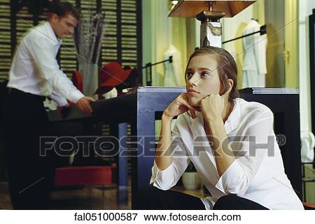 Young woman sitting with hands under chin in clothing store, man looking at table in background View Large Photo Image Stock Photo - Young woman sitting with hands under chin in clothing store, man looking at table in background. Fotosearch
