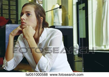 Young woman sitting with hands under chin, looking away in thought View Large Photo Image Stock Photo - Young woman sitting with hands under chin, looking away in thought. Fotosearch