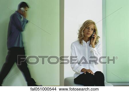Young woman using cell phone, man walking in background, also on cell phone View Large Photo Image Picture - Young woman using cell phone, man walking in background, also on cell phone. Fotosearch