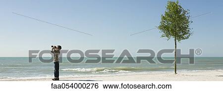 Stock Photograph - Boy standing on beach, looking at sea with binoculars. Fotosearch