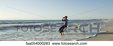Stock Image - Businessman sitting on desk at the beach, looking through binoculars at sea. Fotosearch