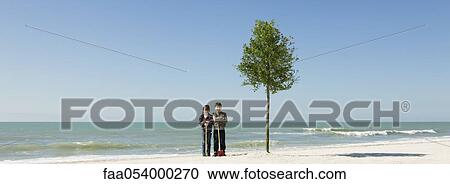 Stock Image - Children standing with shovels beside tree planted on beach. Fotosearch