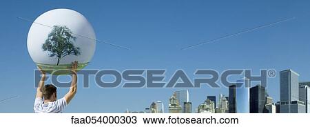 Stock Image - Holding aloft globe displaying image single tree, city skyline in background. Fotosearch