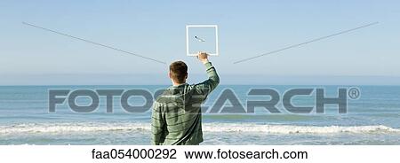 Man on beach holding up picture frame capturing image of gull flying against blue sky View Large Photo Image Stock Image - Man on beach holding up picture frame capturing image of gull flying against blue sky. Fotosearch