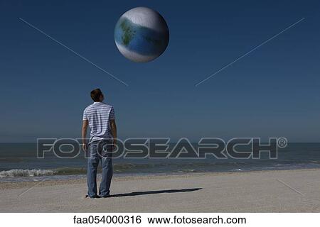 Stock Photograph - Man on beach looking up at alien world orbiting overhead. Fotosearch