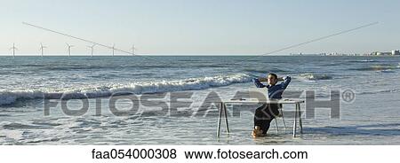 Man relaxing at desk set at water's edge on beach, wind turbines on horizon View Large Photo Image Stock Photo - Man relaxing at desk set at water's edge on beach, wind turbines on horizon. Fotosearch