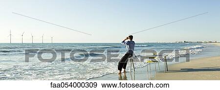 Man sitting on edge of desk at water's edge on beach looking at offshore wind turbines on horizon View Large Photo Image Stock Photo - Man sitting on edge of desk at water's edge on beach looking at offshore wind turbines on horizon. Fotosearch