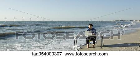 Man working at desk placed at water's edge on beach, wind turbines on horizon View Large Photo Image Stock Photo - Man working at desk placed at water's edge on beach, wind turbines on horizon. Fotosearch