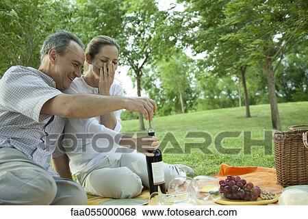 Stock Photo - Mature couple having picnic outdoors, opening bottle of wine. Fotosearch