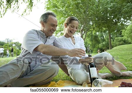 Stock Photo - Mature couple having picnic outdoors, opening bottle of wine. Fotosearch