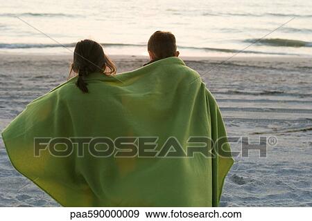 Stock Photo - Children wrapped together in blanket standing on beach observing sunset. Fotosearch