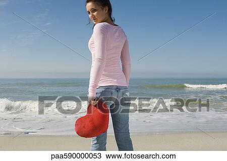 Stock Image - Preteen girl at beach holding stuffed heart-shaped toy, looking over shoulder at camera. Fotosearch
