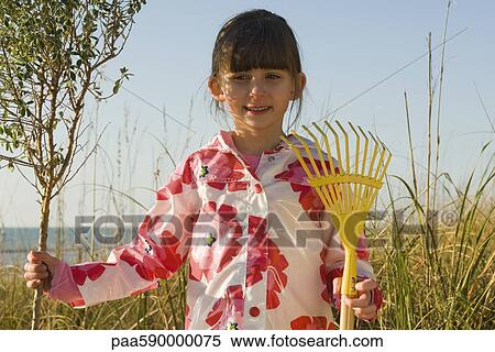 Stock Photography - Young girl with gardening rake standing next to tree sapling. Fotosearch