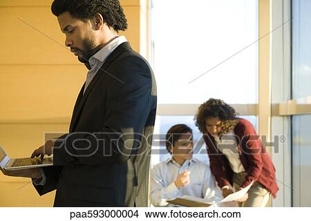 Picture - Executive standing in corridor, using laptop computer, colleagues working in background. Fotosearch