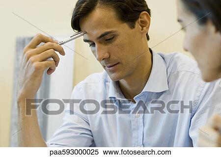 Stock Photography - Man looking down in thought, holding pen against forehead. Fotosearch