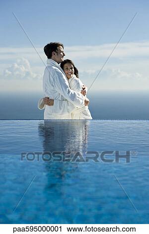 Stock Image - Couple embracing at edge of infinity pool, both wearing bathrobes. Fotosearch