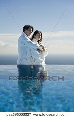Stock Photo - Couple embracing at edge of infinity pool, both wearing bathrobes. Fotosearch