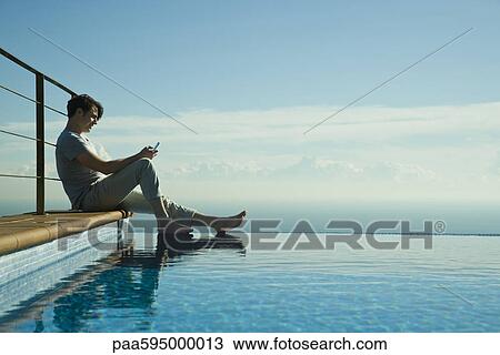 Stock Image - Man sitting on edge of infinity pool, text messaging with cell phone. Fotosearch