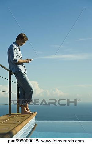 Stock Photography - Man standing beside infinity pool, text messaging with cell phone. Fotosearch