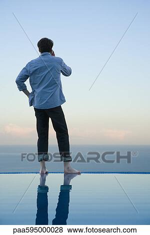 Man standing on edge of infinity pool, talking on cell phone View Large Photo Image Stock Photo - Man standing on edge of infinity pool, talking on cell phone. Fotosearch
