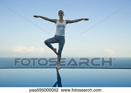 Stock Image - Woman standing in tree pose on edge of infinity pool. Fotosearch