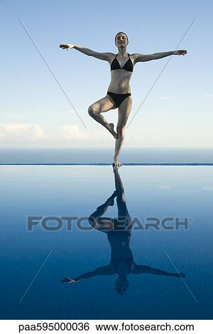 Stock Photograph - Woman standing in tree pose on edge of infinity pool. Fotosearch