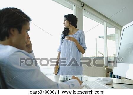 Woman standing with cup of coffee, looking out window, husband working on computer in foreground View Large Photo Image Stock Image - Woman standing with cup of coffee, looking out window, husband working on computer in foreground. Fotosearch