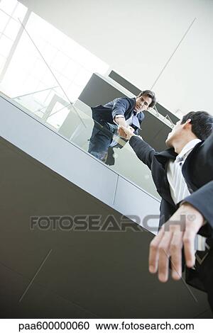 Stock Image - Businessman leaning over balcony, holding on to colleague's hand. Fotosearch