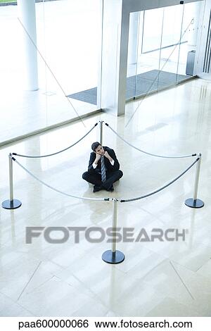 Stock Photograph - Businessman sitting on floor inside roped off area in lobby. Fotosearch