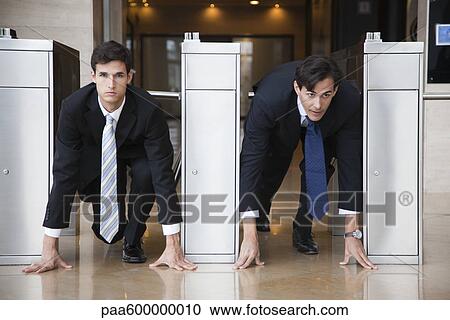 Stock Image - Businessmen crouching in starting position in lobby turnstiles. Fotosearch
