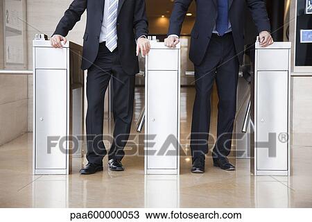 Stock Image - Businessmen walking through turnstiles, cropped. Fotosearch