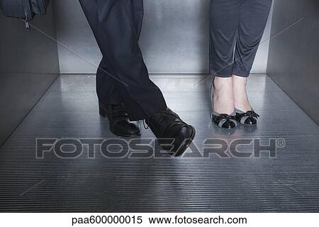 Stock Photography - Man and woman standing in elevator, man moving foot toward woman's foot. Fotosearch