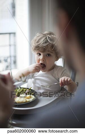 Toddler boy eating meal, looking at parent in foreground View Large Photo Image Stock Image - Toddler boy eating meal, looking at parent in foreground. Fotosearch