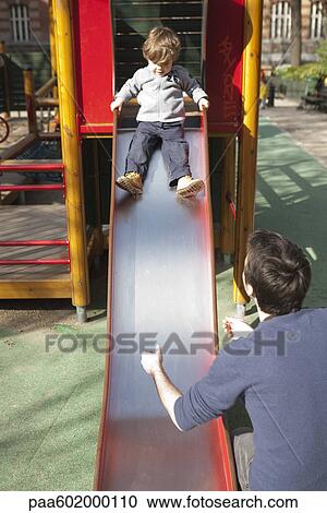 Stock Image - Toddler boy preparing to go down playground slide, father waiting at the bottom. Fotosearch