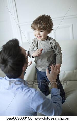 Stock Image - Toddler boy standing on couch, smiling at father. Fotosearch