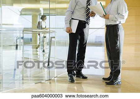 Stock Image - Businessmen discussing document in corridor, cropped. Fotosearch