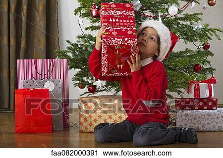 Stock Image - Boy sitting beside Christmas tree, shaking gift. Fotosearch