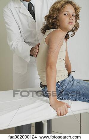 Boy sitting on examination table, doctor examining his back View Large Photo Image Picture - Boy sitting on examination table, doctor examining his back. Fotosearch