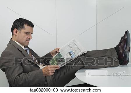 Stock Image - Businessman reading newspaper in office with feet up on desk. Fotosearch