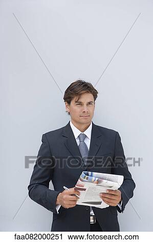 Stock Image - Businessman standing outdoors, holding pen and newspaper. Fotosearch