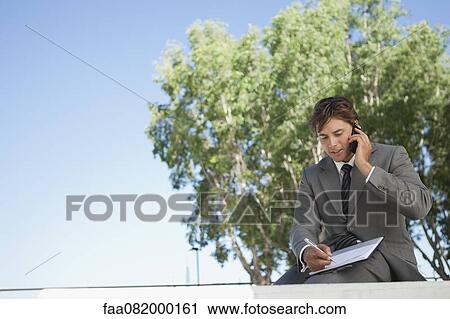 Stock Image - Businessman talking on cell phone and writing on clipboard outdoors. Fotosearch
