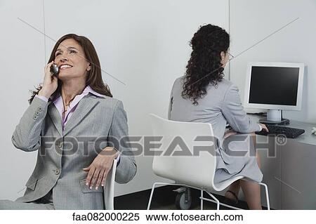 Businesswoman idly chatting on cell phone while colleague works nearby View Large Photo Image Stock Photography - Businesswoman idly chatting on cell phone while colleague works nearby. Fotosearch