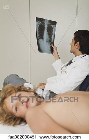 Stock Image - Doctor looking at x-ray, boy lying barechested on examination table. Fotosearch