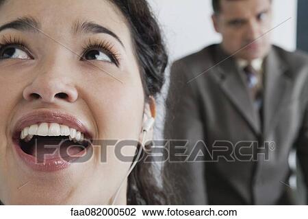 Stock Image - Woman listening to earphones and singing in office, angry boss in background. Fotosearch