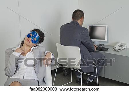 Stock Photo - Woman napping with eye mask in office as colleague works nearby. Fotosearch