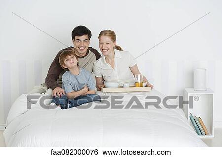 Stock Photograph - Young family sitting in bed, mother holding breakfast tray. Fotosearch