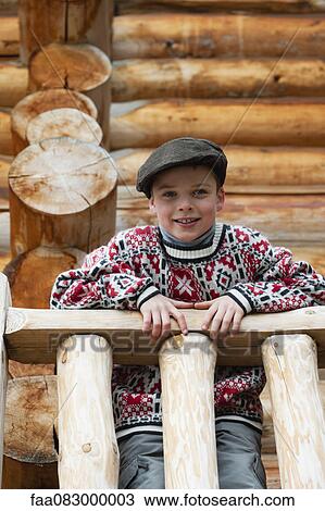 Stock Image - Boy standing on deck of log cabin, looking down at camera, portrait. Fotosearch