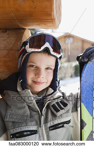 Stock Image - Boy wearing ski goggles and ski jacket and holding snowboard, portrait. Fotosearch